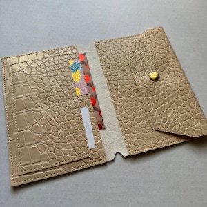 Pode incluir: Uma carteira bege com padrão de pele de crocodilo texturizado e fecho de botão de pressão dourado. O interior contém uma ranhura vertical com um cartão estampado em amarelo, azul e vermelho, ao lado de um cartão retangular branco.