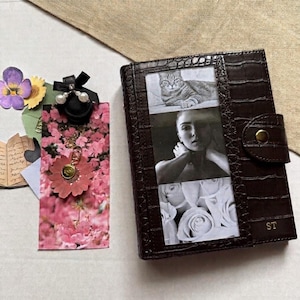 Op de afbeelding: Een donkerbruin notitieboek van leer met krokodillenprint, een drukknoopsluiting en de initialen ST. De kaft bevat een verticale fotostrip met een kat, een persoon en witte rozen. Daarnaast ligt een roze bloemenkaart met een gouden bedel en parels.