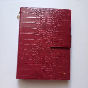 Pode incluir: Um diário de cor bordô com textura de pele de crocodilo. O diário tem uma corrente dourada e um fecho de pressão. As iniciais "JC" estão gravadas em dourado no canto inferior direito.