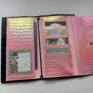May include: A tri-fold wallet featuring a pink iridescent faux-leather exterior with a crocodile-skin texture. The interior contains multiple card slots, a central elastic band, and several decorative inserts, including a plant illustration.