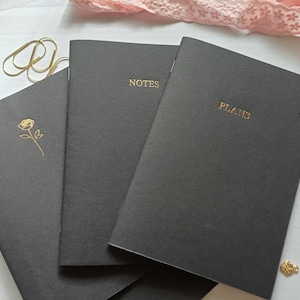 Peut inclure: Trois carnets gris foncé avec des lettres et des accents dorés. L'un porte l'inscription "NOTES", un autre "PLANS", et le troisième présente une illustration de rose dorée. Des trombones et des boucles d'oreilles dorés sont également présents.