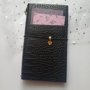 May include: Black faux leather journal with a crocodile skin texture. The journal has a gold heart charm and a black elastic closure. Two decorative cards with gold accents are visible in the front pocket.