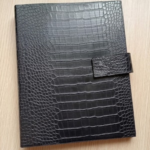May include: Black faux-leather portfolio with a crocodile skin texture. The portfolio has a rectangular shape with a flap closure. The cover is a solid black color, and the texture gives it a luxurious look.