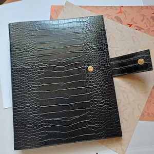 Op de afbeelding: Zwart imitatieleer portfolio met een krokodillenleer textuur. De portfolio heeft een klepsluiting met twee goudkleurige drukknopen. De achtergrond bevat patroonpapier in roze en witte tinten.
