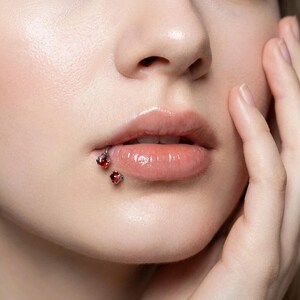 May include: Close-up of a person's lips with two red gemstone lip piercings. The piercings are set in silver-colored metal. The lips are glossy and a natural pink color.