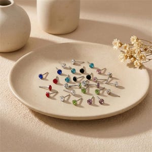 May include: A collection of stud earrings with various colored gemstones, including red, blue, green, and clear, arranged on a beige plate. The earrings have silver posts and are displayed in a well-lit setting.