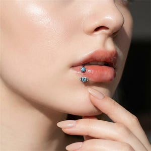 May include: Close-up of a person's lips with two pieces of jewellery. The upper lip has a small, round, blue gem stud. The lower lip has a heart-shaped stud with wings, also with a blue gem.