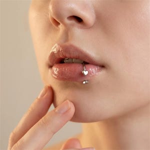 Può includere: Primo piano delle labbra di una persona con un piercing al labbro argentato. Il piercing presenta un piccolo ciondolo a forma di cuore sul labbro superiore e un piccolo borchia sul labbro inferiore. Le labbra sono lucide e di colore rosa chiaro.