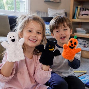 May include: Three crocheted finger puppets featuring a white ghost with a black face, a black bat with yellow accents, and an orange pumpkin with a green stem and black facial features. Each puppet is crafted with a textured yarn stitch pattern.