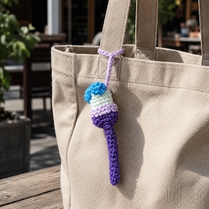 May include: A beige canvas tote bag with a crochet keychain attached to the handle. The keychain features a multi-coloured popsicle design in shades of blue, light green, lavender, and dark purple, suspended by a light purple crochet loop.