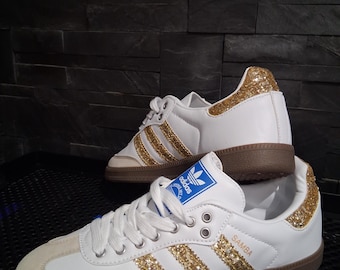 Könnte beinhalten: Weiße Adidas Samba Sneaker mit goldenen Glitzerakzenten auf den drei Streifen und der Ferse. Die Sneaker haben eine braune Sohle und eine Zehenkappe aus Wildleder. Das Wort "Samba" ist auf der Seite aufgedruckt.