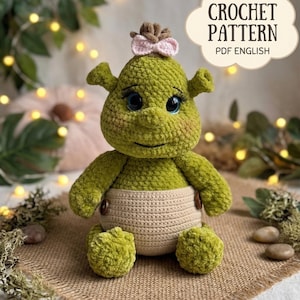 Può includere: Bambola all'uncinetto verde fatta a mano con pannolino marrone chiaro e fiocco rosa. La bambola ha gli occhi blu ed è seduta su una superficie di iuta. Il testo "CROCHET PATTERN PDF ENGLISH" è incluso.