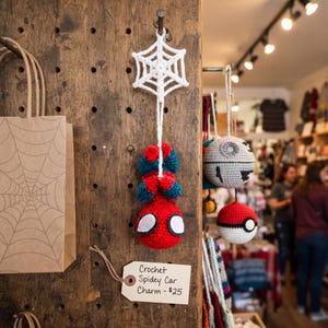 May include: A handmade crochet hanging ornament featuring a red and blue character with large white eyes, suspended from a white crocheted spider web. A rectangular paper tag attached to the display board reads Crochet Spidey Car Charm.
