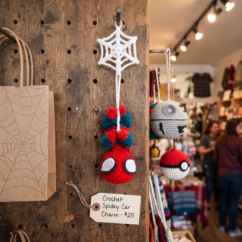 May include: A handmade crochet hanging ornament featuring a red and blue character with large white eyes, suspended from a white crocheted spider web. A rectangular paper tag attached to the display board reads Crochet Spidey Car Charm.