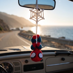 May include: A crocheted hanging ornament featuring a red and blue superhero figure suspended from a white crocheted spider web. The figure has a round red head with large white eyes, a red and blue body, and is attached to a white string.