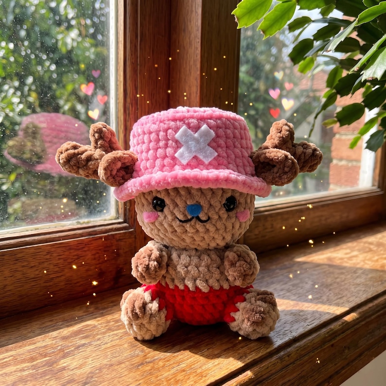 May include: A handmade crochet plush toy of a cartoon character. The plush has a pink hat with a white cross, brown antlers, and a tan body with red shorts. The toy has black eyes, pink cheeks, and a blue nose.