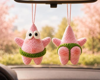 May include: Two pink crochet star-shaped ornaments hanging from a rearview mirror. One shows the front with large white eyes and a green waistband, while the other shows the back with a green thong-style garment over the pink textured surface.