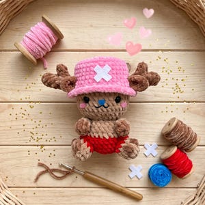 May include: A handmade crochet plush toy of a cartoon character, wearing a pink hat with a white cross, and red shorts. The toy is surrounded by crafting supplies, including spools of yarn, a crochet hook, and decorative hearts.