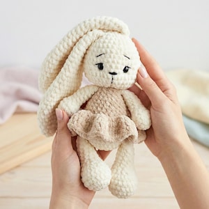 Puede incluir: Muñeca de conejo de ganchillo hecha a mano en crema y marrón. El conejo tiene orejas largas y caídas, una carita con detalles cosidos en negro y un vestido marrón. La muñeca es suave y afelpada, perfecta para un juguete infantil o decoración de guardería.