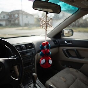 May include: A crocheted hanging ornament featuring a red and blue superhero figure suspended from a white yarn spiderweb. The figure has a round red head with large white eyes and is attached to the web by a single white string, dangling from a car mirror.