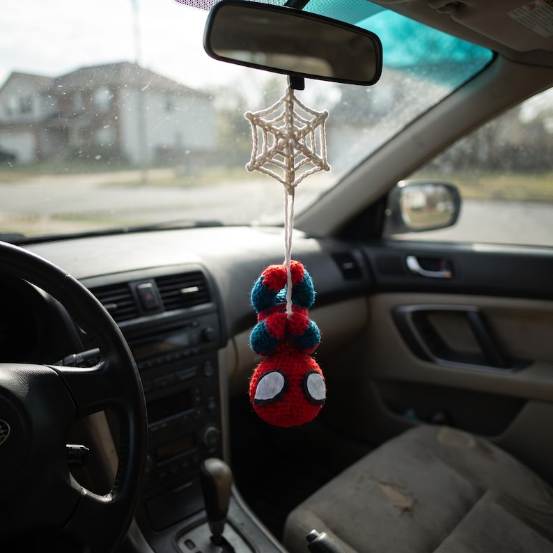 May include: A crocheted hanging ornament featuring a red and blue superhero figure suspended from a white yarn spiderweb. The figure has a round red head with large white eyes and is attached to the web by a single white string, dangling from a car mirror.