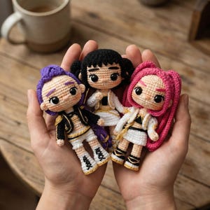 May include: Three handmade crochet dolls with different hair colors: purple, black, and pink. Each doll wears a unique outfit with black and gold accents. The dolls are held in open hands.