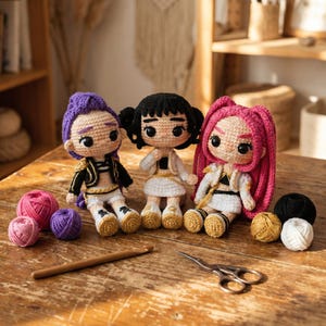 May include: Three handmade crochet dolls with different hair colors: purple, black, and pink. Each doll is wearing a unique outfit with gold accents. Yarn balls in pink, purple, black, gold, and white are arranged near the dolls, along with a crochet hook and scissors.