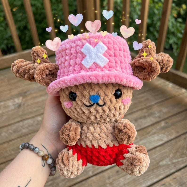 May include: A handmade plush toy of a cartoon character, featuring a pink hat with a white cross, brown antlers, and red shorts. The plush is light brown with a blue nose and pink cheeks. Decorative hearts and stars are added to the image.