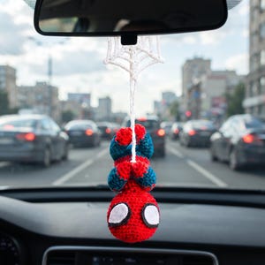May include: A crocheted hanging ornament featuring a red and blue character with large white eyes, suspended from a white crocheted spider web design. The figure is positioned upside down, attached by a white string to a car rearview mirror.