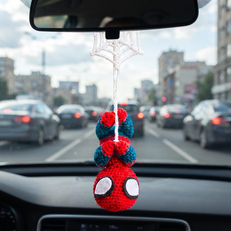 May include: A crocheted hanging ornament featuring a red and blue character with large white eyes, suspended from a white crocheted spider web design. The figure is positioned upside down, attached by a white string to a car rearview mirror.