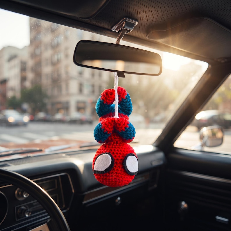 May include: A crocheted hanging ornament featuring a red and blue character with large white eyes, suspended by a white string from a vehicle rearview mirror. The figure is depicted in a downward-facing position with its arms folded near its head.