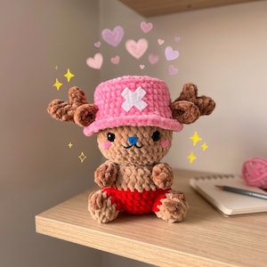 May include: A handmade plush toy of a cartoon character, featuring a pink hat with a white cross, brown antlers, and red shorts. The toy is sitting on a wooden shelf, with hearts and sparkles in the background.