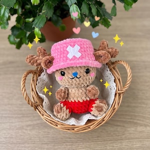 May include: A handmade plush toy of a cartoon character, featuring a pink hat with a white cross, brown antlers, and red shorts. The toy is nestled in a wicker basket with a lace doily, surrounded by decorative hearts and stars.