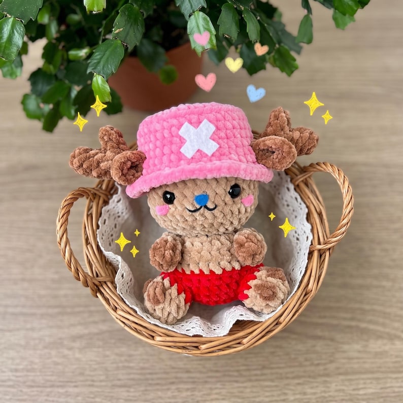 May include: A handmade plush toy of a cartoon character, featuring a pink hat with a white cross, brown antlers, and red shorts. The toy is nestled in a wicker basket with a lace doily, surrounded by decorative hearts and stars.