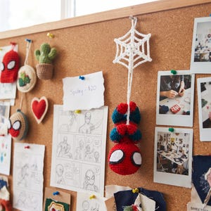 May include: A crocheted Spider-Man figure hanging upside down from a white crocheted spider web. The figure is red and blue with large white eyes. The web is attached to a cork board featuring various sketches, photos, and other small items.