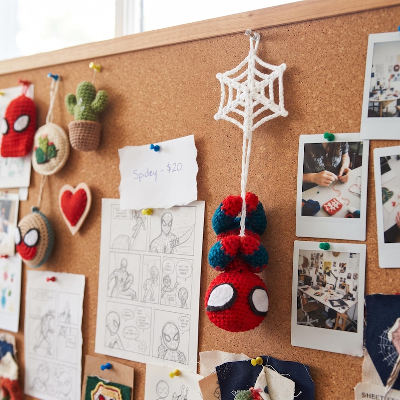 May include: A crocheted Spider-Man figure hanging upside down from a white crocheted spider web. The figure is red and blue with large white eyes. The web is attached to a cork board featuring various sketches, photos, and other small items.