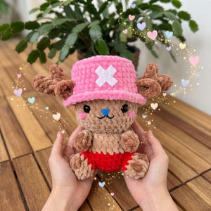 May include: A handmade crochet plush toy of a cartoon character. The toy has a pink hat with a white cross, brown antlers, and red shorts. The plush is held in hands, with a background of a plant and wooden table.