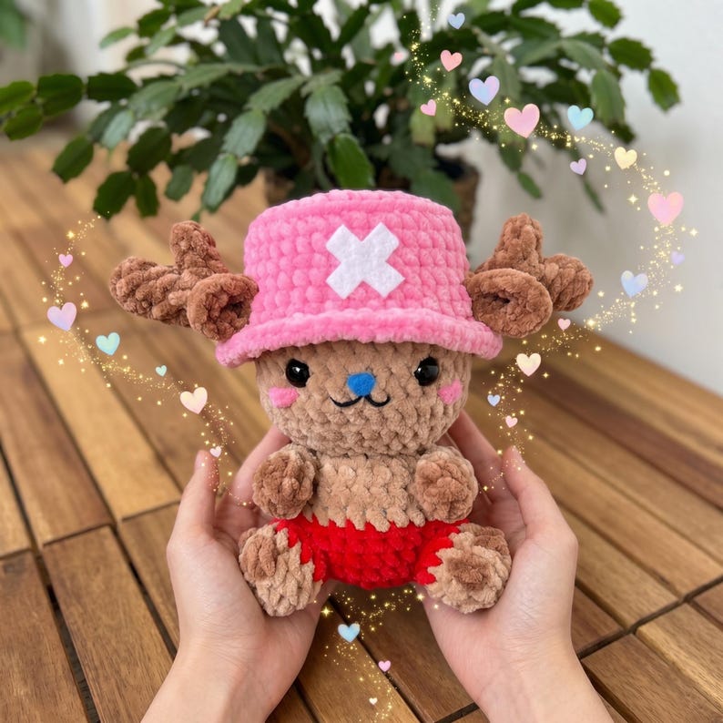 May include: A handmade crochet plush toy of a cartoon character. The toy has a pink hat with a white cross, brown antlers, and red shorts. The plush is held in hands, with a background of a plant and wooden table.