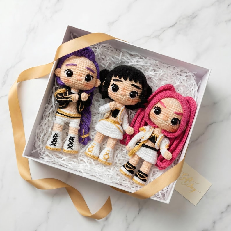 May include: Three handmade crochet dolls with different hairstyles and outfits are presented in a white gift box with gold ribbon. The dolls have black, purple, and pink hair, and wear white, gold, and black outfits. The dolls are approximately 6 inches tall.