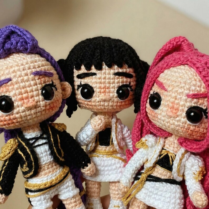 May include: Three handmade crochet dolls with different hair colors: purple, black, and pink. Each doll wears a white outfit with gold accents and a jacket. The dolls have large, black eyes and rosy cheeks, giving them a cute appearance.