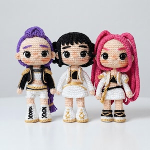 Puede incluir: Tres muñecas de ganchillo hechas a mano con cabello morado, negro y rosa. Cada muñeca viste un atuendo blanco con detalles dorados, una chaqueta negra y botas blancas. Las muñecas tienen ojos grandes y expresivos y están una al lado de la otra.