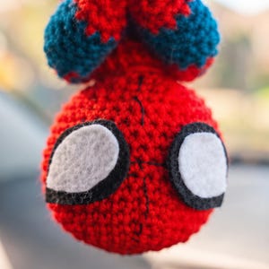 May include: A round, crocheted yarn ornament featuring a red and blue color scheme. The object has two large, white, felt-like eye patches outlined in black thread, with black stitching details running vertically down the center of the red face.