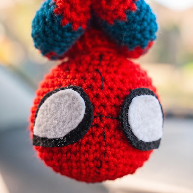 May include: A round, crocheted yarn ornament featuring a red and blue color scheme. The object has two large, white, felt-like eye patches outlined in black thread, with black stitching details running vertically down the center of the red face.