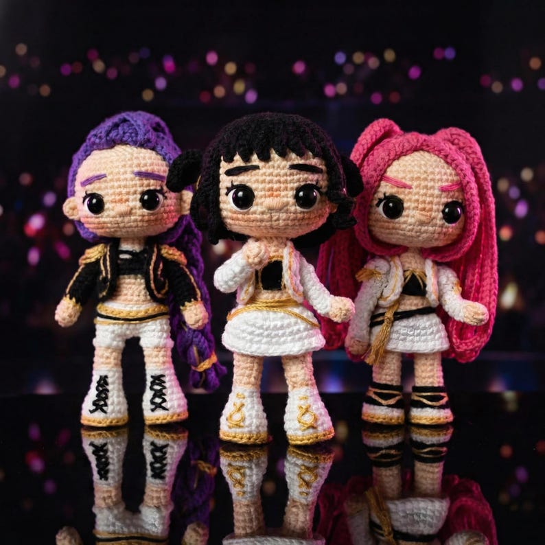 May include: Three handmade crochet dolls with different hair colors: purple, black, and pink. Each doll wears a white skirt, jacket, and boots with gold accents. The dolls have large, expressive eyes and are standing on a reflective surface.