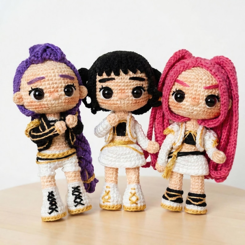 May include: Three handmade crochet dolls with detailed features. Each doll has a unique hairstyle in purple, black, and pink, and wears a white outfit with gold and black accents. The dolls have large, expressive eyes and are standing on a light-colored surface.