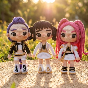 May include: Three handmade crochet dolls with different hairstyles and outfits. The dolls have black eyes and are standing on a textured surface. The dolls are wearing white skirts, jackets, and boots with gold and black accents. One doll has purple hair, one has black hair, and one has pink hair.