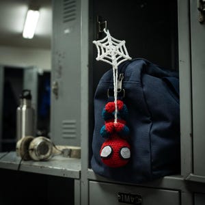 May include: A small, handmade crochet figure of a red and blue superhero hanging upside down from a white crocheted spiderweb. The figure is attached to the zipper of a dark navy blue canvas duffel bag, which is resting inside a gray metal locker.