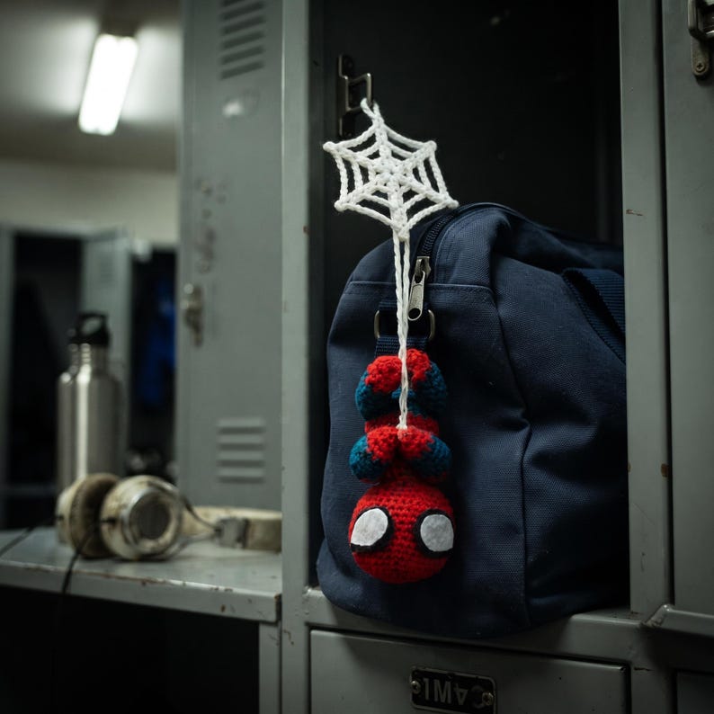 May include: A small, handmade crochet figure of a red and blue superhero hanging upside down from a white crocheted spiderweb. The figure is attached to the zipper of a dark navy blue canvas duffel bag, which is resting inside a gray metal locker.