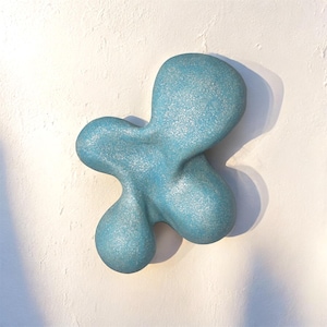 Pode incluir: Escultura de parede abstrata azul claro com uma superfície texturizada e brilhante. A forma orgânica tem múltiplos lóbulos arredondados, criando um design único e moderno. A escultura é montada em uma parede branca texturizada.