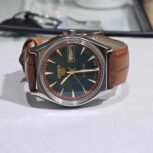 Peut inclure: Une montre-bracelet automatique Seiko 5 dotée d'un cadran texturé vert foncé avec des aiguilles et des index dorés. La montre possède un boîtier en acier inoxydable argenté et un bracelet en cuir marron avec un grain et des coutures blanches.